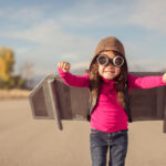 A young girl is wearing goggles and a flight cap and is ready to take to the sky. She has made her own jet pack and is ready to fly away to her dreams. The girl is standing on a road in rural Utah, USA. Girls have big ideas too.